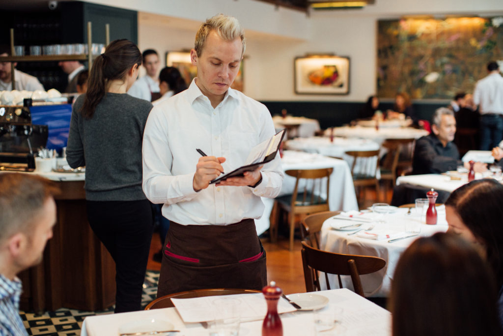 Photo of a server in a white button down shirt taking an order tableside. 