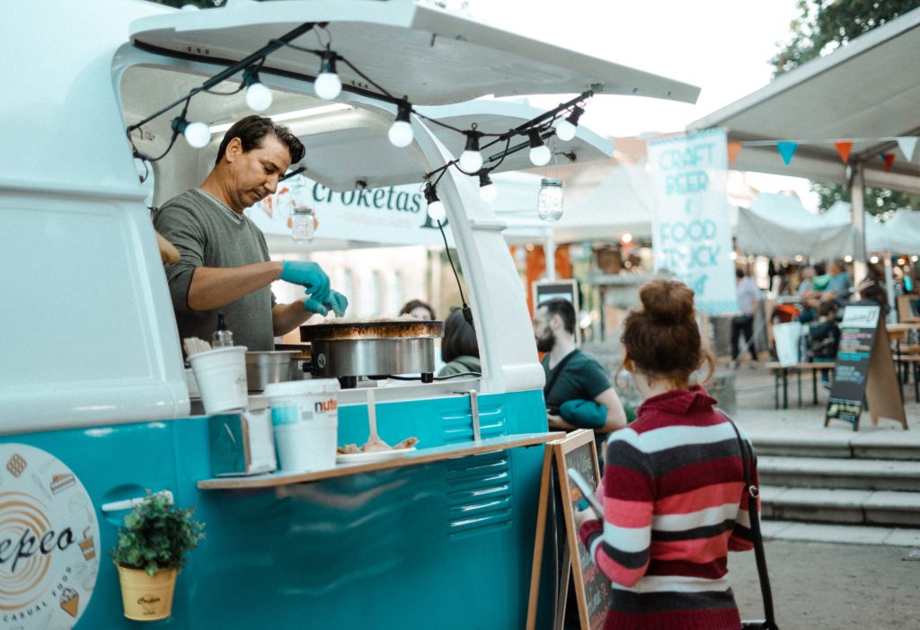 Photo of a man in a turquoise and white food truck making food with a woman in a striped sweater waiting. 