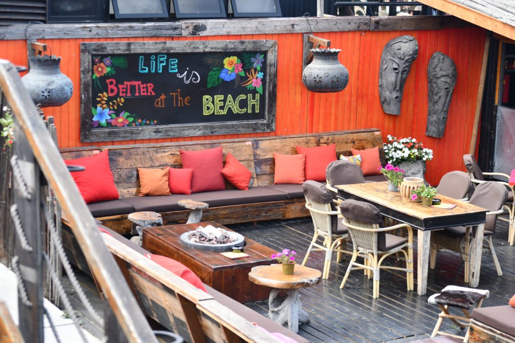 Photograph of a restaurant balcony with outdoor seating. A large chalkboard can be seen on one wall with "Life is Better at the Beach" printed in colorful chalk letters with flowers. 