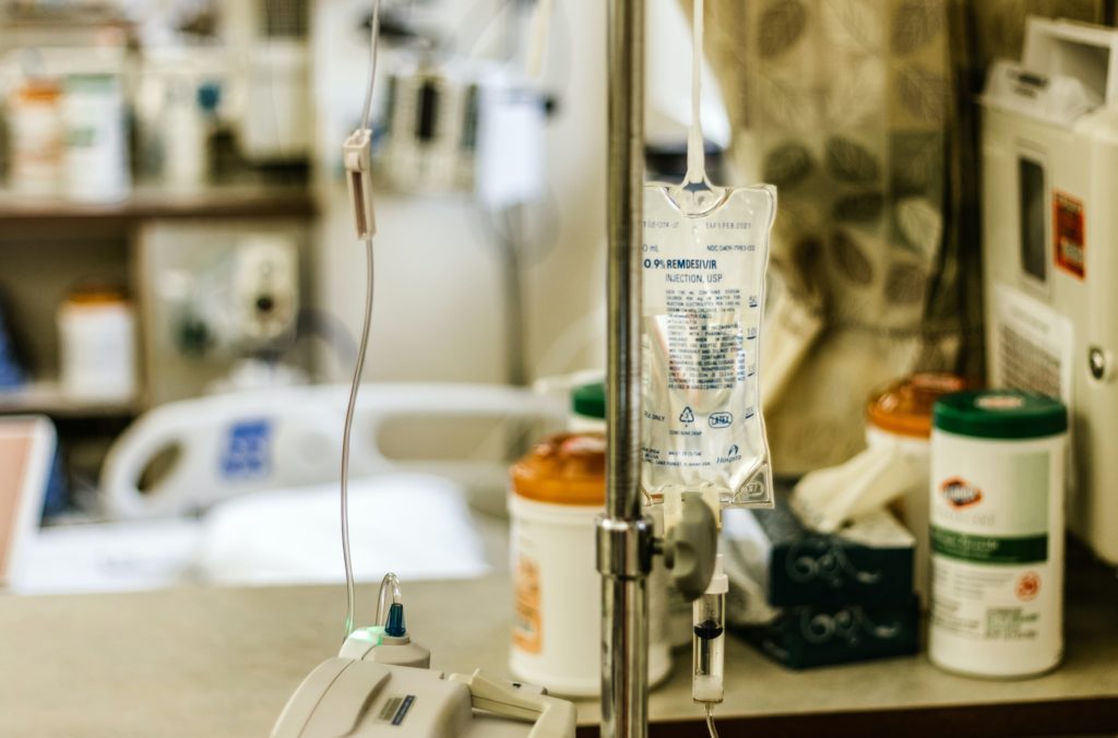 Photo of an IV pole with a bag labeled "Remdesivir" hanging with a blurred hospital setting in the background. Image by Dimitri Karastelev on Unsplash.  