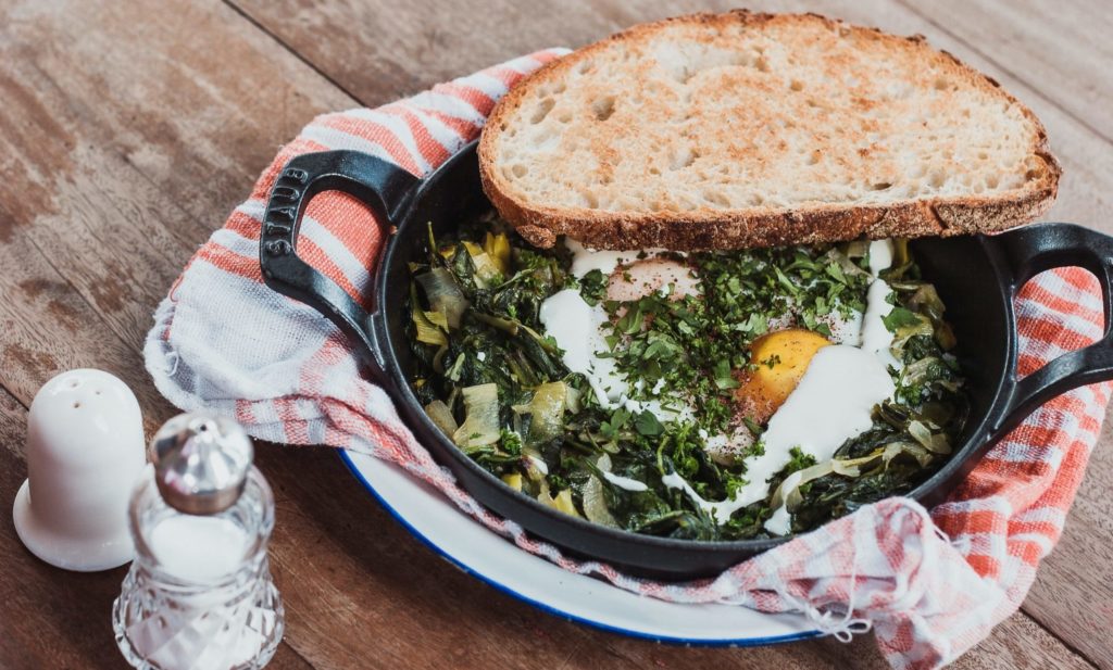 Photograph of a small cast iron skillet with greens, eggs, and a slice of toast sitting on a plate with a cloth napkin. 
