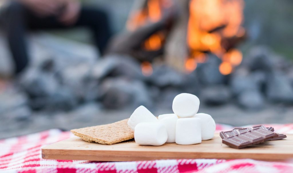 Photograph with a campfire in the background and a wooden board on a red checkered table cloth holding graham crackers, marshmallows, and pieces of chocolate.