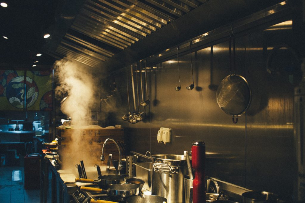 Photo of a steamy industrial kitchen with soup ladles hanging on a steel wall and several pans on a stovetop. Photo credit: Francisco Suarez via Unsplash.  