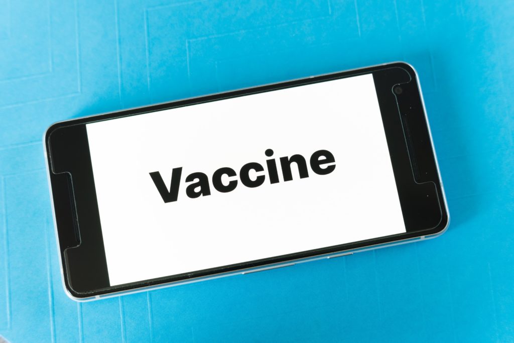 Photo of a mobile phone on a blue background with the word "Vaccine" on the display. Photo credit: Markus Winkler via Unsplash
